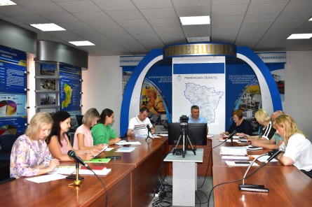 На фото — робоче засідання за участю представників Рівненської області. Учасники сидять за столами, працюють із документами та обговорюють робочі питання. У приміщенні встановлені мікрофони та камера для запису заходу, на задньому плані — інформаційні стенди з матеріалами про Рівненщину.