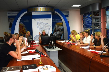 На фото — робоче засідання за участю представників Рівненщини. Учасники сидять за столами з документами та матеріалами, частина з них підняла руки під час голосування. У центрі приміщення встановлені монітор і камера для онлайн-зв’язку, на задньому плані — інформаційні стенди та карта Рівненської області.