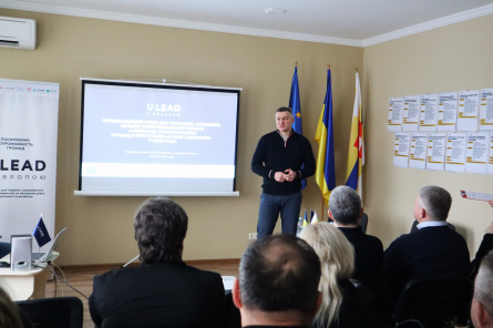 Біля мультимедійного екрану та прапорів Рівненщини, Євросоюзу та України стоїть заступник голови Рівненської ОДА Олександр Кохан. Учасники зустрічі слухають, про що він говорить