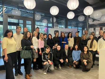 На фото — група людей, які зібралися для спільного знімка в сучасному приміщенні, схожому на простір для заходів або бізнес-зустрічей. Усього близько двох десятків учасників: частина стоїть, а кілька людей присіли попереду.

У центрі композиції — жінка в яскравому синьому піджаку, поруч із нею інші учасники, одягнені переважно у діловому або повсякденно-діловому стилі. Люди виглядають зосередженими та спокійними, більшість дивиться в камеру.

Позаду розміщений стенд із написом «ДІЯ.БІЗНЕС», а праворуч видно інформаційні панелі з логотипами партнерів. Приміщення має великі скляні стіни, через які видно вулицю, а зі стелі звисають круглі білі світильники, що створюють м’яке освітлення.

Загалом фото передає атмосферу ділової події, зустрічі або навчального заходу для підприємців чи учасників бізнес-ініціатив.