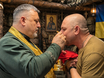 На фото військовий капелан, отець Василь Лозинський причащає військовго в бліндажі, у зоні виконання бойового завдання бригадою. На задньому плані ікона Ісуса Христа.