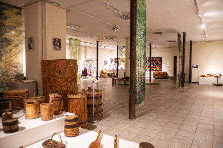 На фото зображено просторий виставковий зал, схожий на музей або культурний центр. Приміщення світле, з високою стелею, колонами та рівномірним освітленням.

На передньому плані розташовані експонати з дерева — різноманітні бочки, відра, посуд та інші предмети побуту, ймовірно традиційні або ремісничі вироби. Вони виставлені на білих підставках для огляду відвідувачами.

У глибині залу видно інші експозиції: народний одяг на манекенах (вишиванки та традиційні костюми), а також декоративні килими з яскравими орнаментами на стінах. Також є інформаційні стенди та вітрини з дрібнішими предметами.

Загалом атмосфера спокійна й пізнавальна — це місце, присвячене українській культурі, традиціям і народному мистецтву.