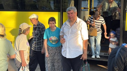 На фото люди виходять із жовтого автобуса. Серед пасажирів — чоловіки, жінки та маленька дитина, яку тримають за руку. Вони мають із собою сумки та речі, що створює враження прибуття або евакуації.