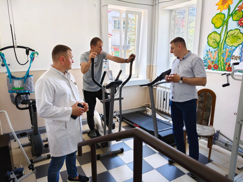 На фото у світлому кабінеті медичного закладу стоїть сучасний тренажер на якому займається ветеран. Біля нього стоять двоє чоловіків: один у білому халаті, очевидно лікар та інший, який дивиться, як проходить заняття спортом