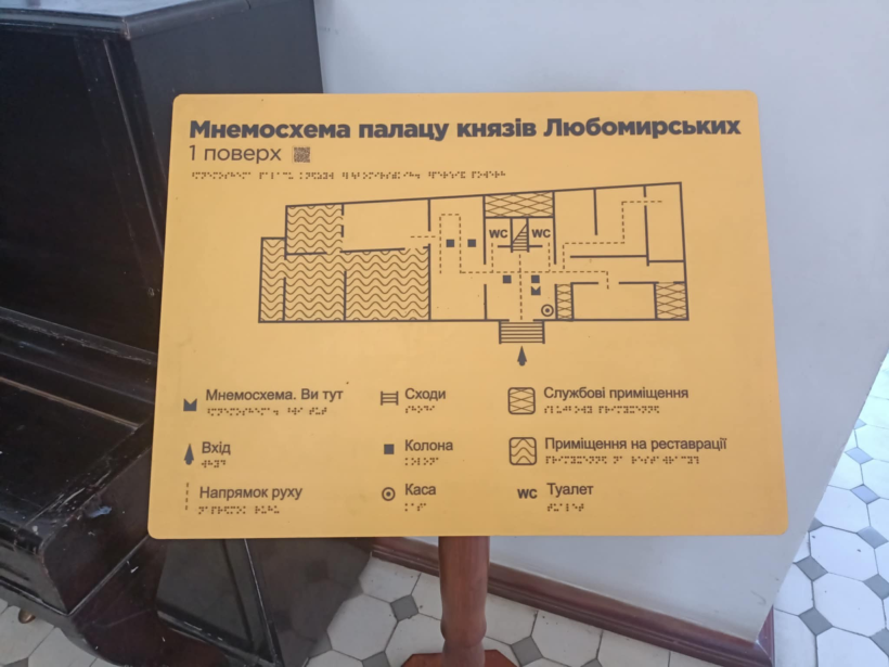 Інформаційна дошка жовтого кольору на якому розміщена інформація про замок українською мовою та шрифтом Брайля