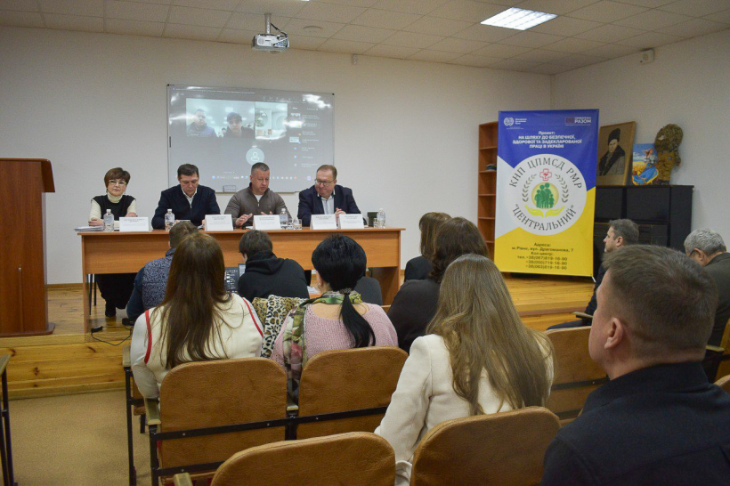На сцені за столом сидять троє чоловіків та жінка. В залі сидять ще чоловіки та жінки. На столі перед дспікерами стоять таблички з їхніми іменами та посадами. Праворуч банер, де на жовто-блакитному тлі напис ЦНП ЦСПМСД РОР "Центральний". Позаду спікерів екран із презентацією