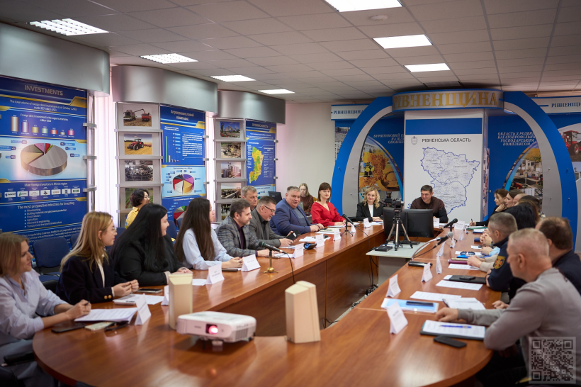 У залі засідань відбувається робоча нарада: учасники сидять за великим столом, обговорюють питання та працюють із документами. На стінах — інформаційні стенди про Рівненську область, у центрі — техніка для презентацій і відеозапису.