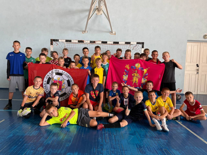Група дітей стоять у спортзалі та тримають у руках прапор із гербом Запоріжжя та прапор Запорізької обласної асоціації футболу.