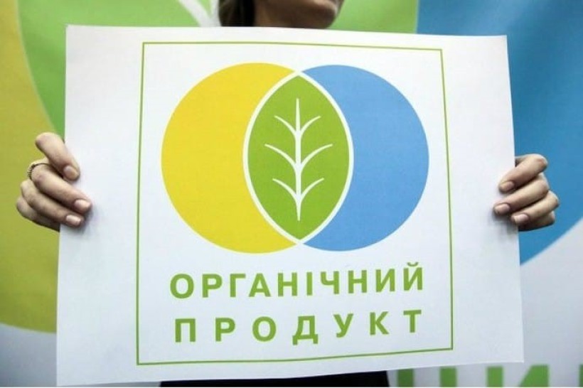 Людина тримає в руках плакат з зображенням жовтого та синього кіл, які перетинаються. У частині, що перетинається, - схематичне зображення ростка рослини. Під зображенням текст "Органічний продукт".