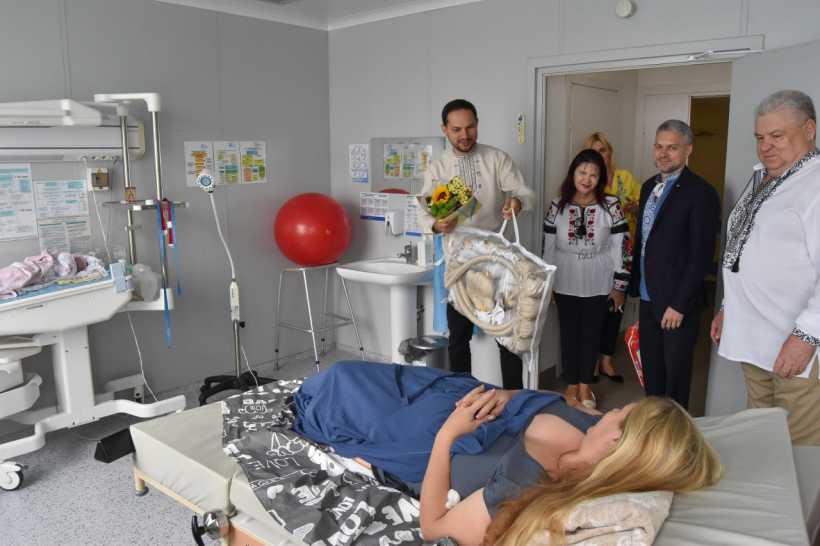 Начальник рівненської ОВА Олександр Коваль заходить у палату, де на ліжку лежить жінка, яка нещодавно народила. Олександр Коваль дарує їй полдарунок у пакеті та букет квітів, він святково одягнутий у вишиванку та посміхається. Разом з ним у палаті ще завідувачка перинатального центру та заступник голови Рівненської ОДА Ігор Павленко