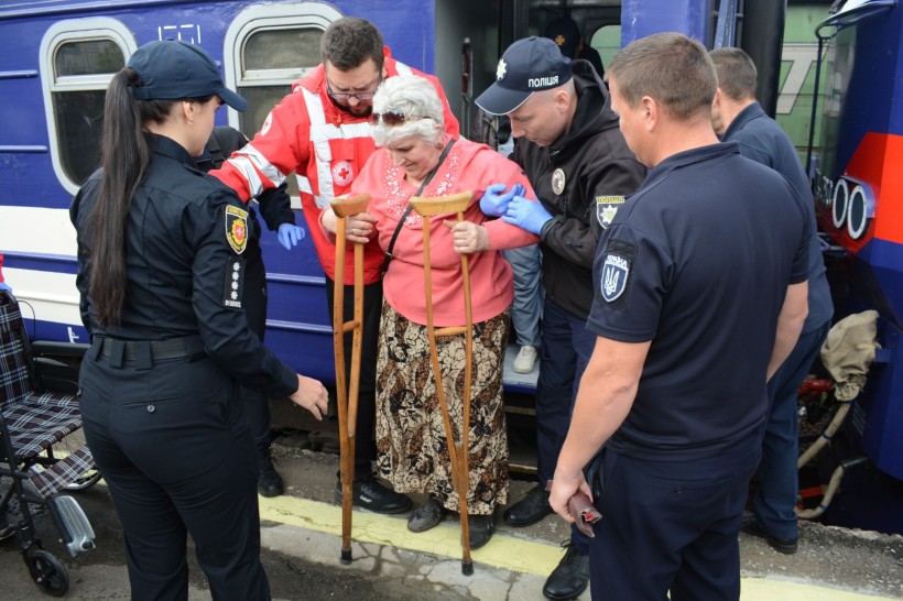 З вагона поїзда на милицях виходить старша сива жінка у рожевій куртці. Їй допомагають зійти на перон молоді чоловіки у формах ДСНС, поліції та представник Червоного хреста