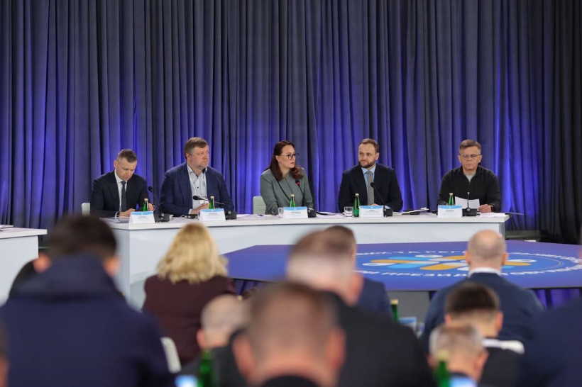 За білим столом сидять представники Уряду та народні депутати. В залі сидять люди та слухають виступи спікерів