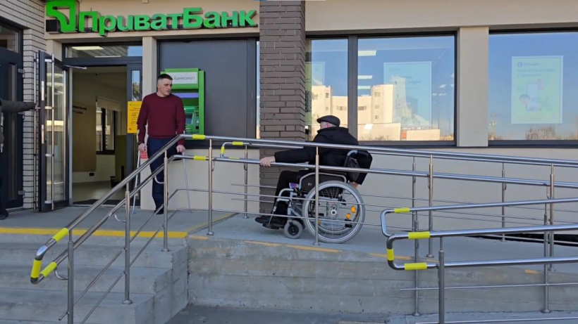 На фото зображено будівлю до якої облаштовано пандус по якому їде чоловік на кріслі колісному. Ще один чоловік без ноги із милицями у руках стоїть біля входу  у цю будівлю, за його спиною банкомат Приватбанку