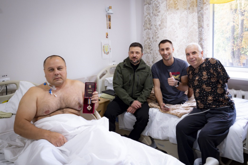 На фото зображено лікарняну палату. Ліворуч — поранений військовий у ліжку, він тримає у руках державну нагороду в червоному футлярі. Поруч із ним сидять троє чоловіків, один з яких Президент України Володимир Зеленський. Двоє чоловіків у повсякденному одязі, один із них показує жест «лайк». Атмосфера світла й щира: поранений воїн отримав відзнаку за службу та сфотографувався разом із відвідувачами й побратимами.