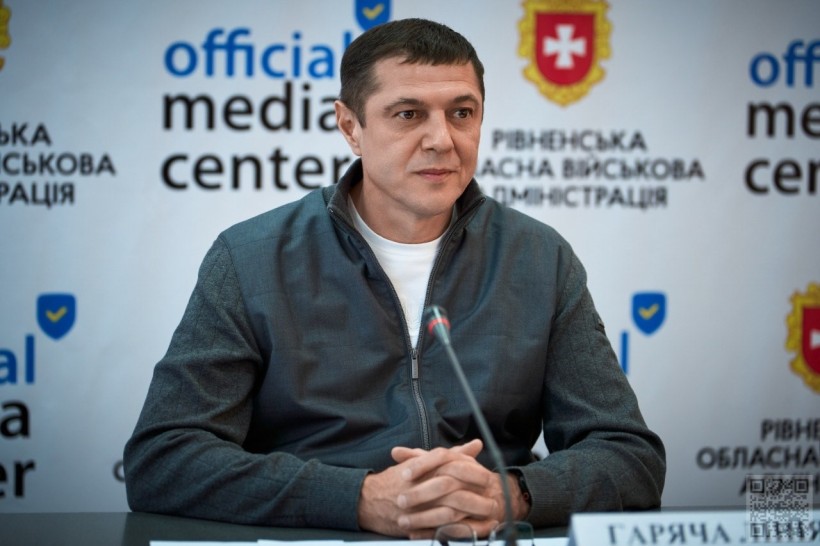 Сергій Подолін сидить у залі за дерев'яним столом, перед ним мікрофон, позаду банер із написом: Рівненська  обласна державна адміністрація