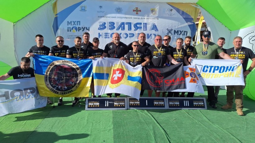 Спортсмени-ветерани Рівненщини на фоні банера Всеукраїнського ветеранського турніру «Звитяга Нескорених»  тримають в руках прапори Рівненської області, спортивного простору "Сила", з написами "Стронг ветеран Рівне", "Розвідувальна рота. Без права на славу. 54 ОМБр. Група Тайсона"