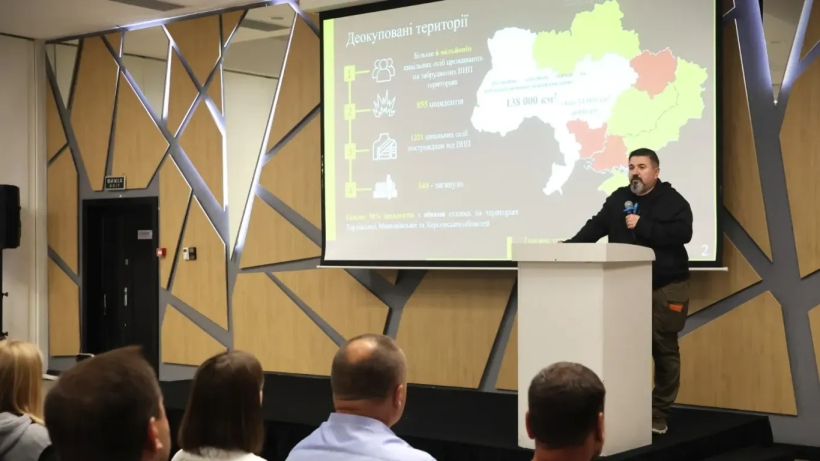 Спікер звертається до присутніх під час форуму «Особливості інформування населення про ризики, пов'язані з мінами та іншими вибухонебезпечними предметами». Він стоїть на підвищенні та говорить у мікрофон. Позаду за ним на моніторі виведено презентацію. У залі сидять слухачі та уважно слухають спікера