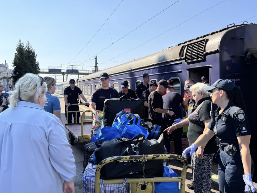 На пероні Рівненського вокзалу біля вагона евакуаційного поїзда стоять до десяти осіб. серед них працівники поліції, ДСНС, інших служб та внутрішньо переміщені особи. У центрі великий возик для перевезення вантажів, на якому зложено багато сумок