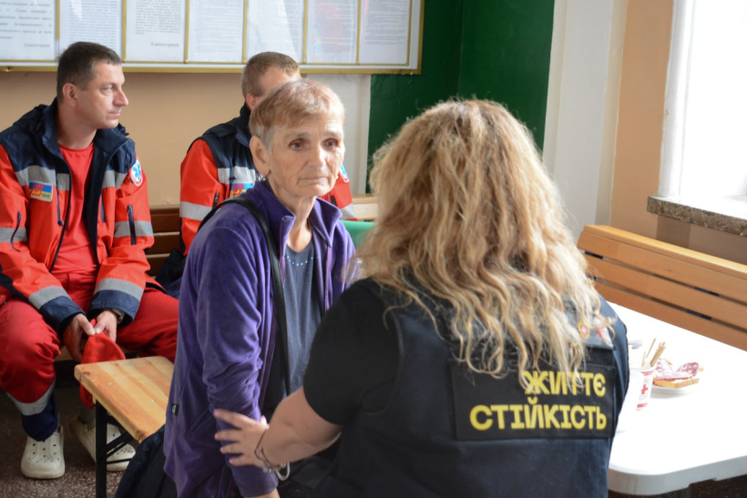 Працівниця центру життєстійкості тримає за руку старшу жінку і говорить з нею. Очевидно, що це психологиня. Жінка сидить на лавці, на її обличчі розгубленість та сум