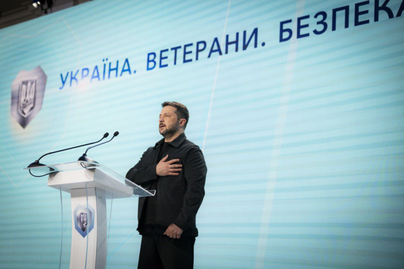 Президент України Володимир Зеленський на сцені під час виконання Державного гімну України. Він стоїть біля трибуни та тримає руку на сарці. На задньому плані банер із написом: Україна.Ветерани. Безпека