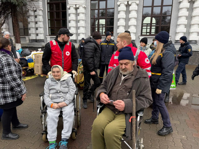 На фото — сцена на відкритому просторі біля будівлі залізничного вокзалу. У центрі уваги — двоє людей старшого віку на кріслах колісних: жінка у світлому спортивному одязі та чоловік у темній куртці й шапці, з палицею в руках.
Навколо них — волонтери Товариства Червоного Хреста у червоних жилетах, а також представники екстрених і правоохоронних служб. Поруч видно коробки з гуманітарною допомогою та особисті речі. Атмосфера фото передає момент організованої підтримки й турботи про людей, які потребують допомоги, у публічному місці.