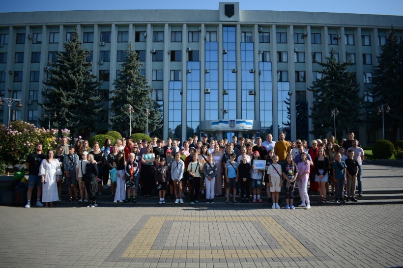Загальне фото на якому на фоні Рівненської облдержадміністрації стоїть група дітей, які їдуть на відпочинок, їх батьків та представників місцевої влади