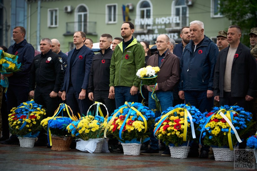 На майдані Незалежності стоять вишикувані учасники урочистостей. В центрі - начальник Рівненської ОВА олександр Коваль, народні депутати, представники органів сісцевого самоврядування, силовики. В них на одязі на грудях прикріплені червоні маки - символи перемоги у Другій світовій війні.  перед ними стоять великі корзини квітів із жовто-блакитними стрічками