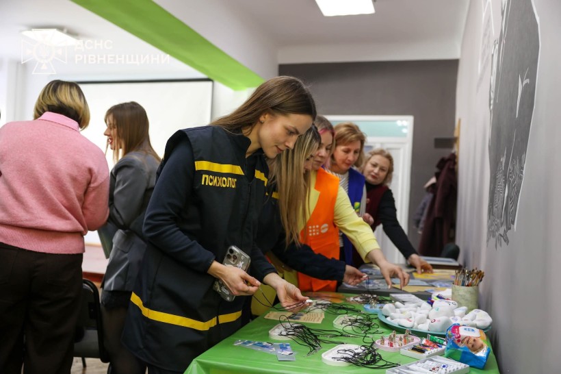 На фото зображено робочу зустріч або презентацію в приміщенні навчального чи адміністративного закладу. Кілька жінок оглядають інформаційні та навчальні матеріали, розкладені на столі.
У центрі уваги — жінка у формі ДСНС із написом «Психолог», яка уважно розглядає один із представлених матеріалів. Поруч інші учасниці заходу знайомляться з експозицією, обговорюють побачене та вибирають предмети зі столу.
На столі розміщені різноманітні роздаткові матеріали, канцелярія, бейджі, інформаційні картки та тематичні предмети, ймовірно для тренінгу або навчального заходу.
У верхньому лівому куті видно логотип ДСНС Рівненщини, що свідчить про причетність служби до організації події. Атмосфера фото ділова, доброзичлива та спрямована на навчання, підтримку чи психологічну роботу з людьми.