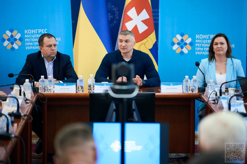 У центрі за столом сидить заступник голови Рівненської ОДА Олександр Кохан, який виступає перед учасниками та жестикулює під час промови. Праворуч і ліворуч від нього — ще двоє учасників зустрічі. Перед кожним розміщені мікрофони, пляшки з водою та іменні таблички.

На задньому плані видно Державний Прапор України та прапор із символікою регіону, а також брендований синій інформаційний банер із написом про Конгрес місцевих та регіональних влад при Президентові України.

На передньому плані — екрани системи відеоконференції, що свідчить про проведення заходу у форматі офлайн/онлайн. Загалом фото передає атмосферу офіційної робочої зустрічі та професійного обговорення.