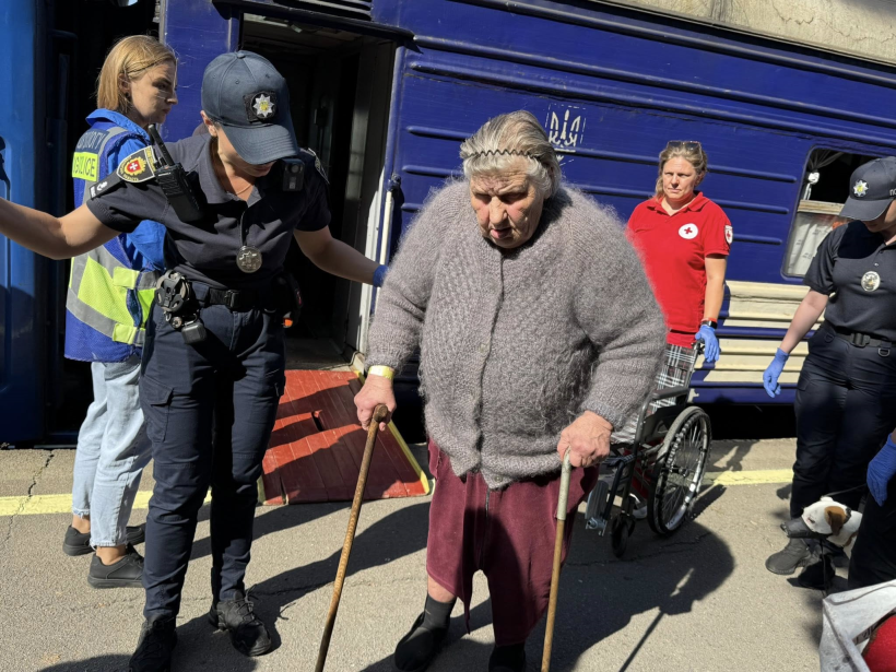 Жінка похилого віку із двома милицями виходить із вагона поїзда. Їй допомагають працівники поліції та Червоного хреста