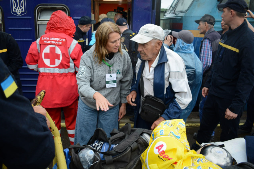 З евакуаційного поїзду виходять люди: старшого чоловіка зустрічають представники Червоного хреста та інших державних установ. На передньому плані багато сумок і жінка з бейджиком на грудях щось пояснює старенькому чоловікові із сивим волоссям у білій кепці та спортивній куртці. Він її уважно слухає. Навколо багато людей.