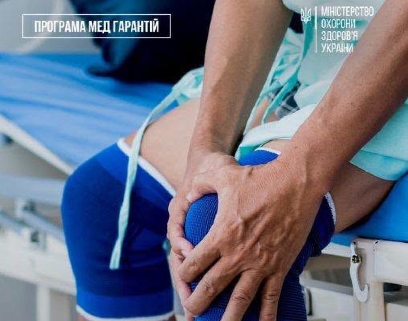 На фото зображена людина, яка сидить на лікарняному ліжку та тримається за коліно. На коліні видно медичний бандаж. У верхній частині зображення розміщені логотипи «Програма медичних гарантій» та Міністерства охорони здоров’я України.