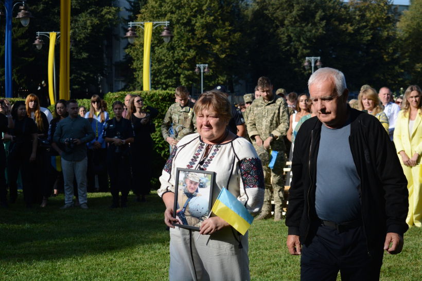 Чоловік та жінка старшого віку ідуть галявиною біля облдержадміністрації. Жінка одягнута у вишиванку, вона тримає у руках портрет загиблого сина та маленький жовто-блакитний прапорець