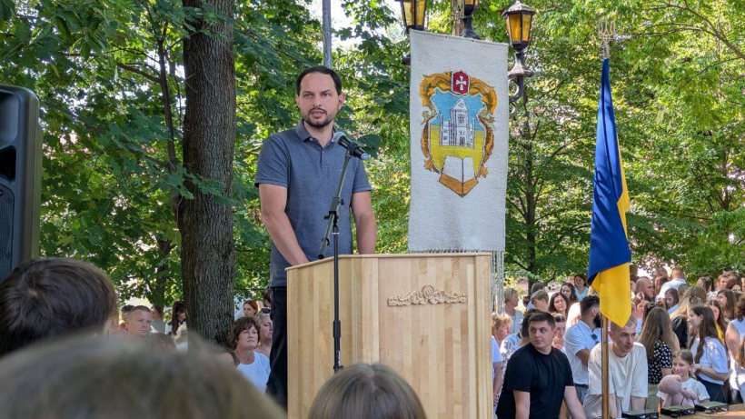 Начальник Рівненської ОВА Олександр Коваль стоїть за трибуною перед студентами звертається до студентів, викладачів та батьків. Трибуна знаходиться на вулиці біля університету, за ним зеленіє парк