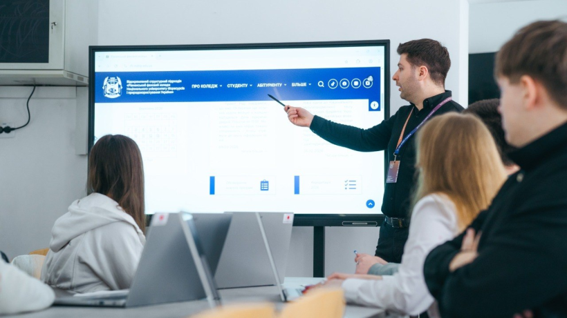 Фото ілюстративне -  зображено навчальний процес у сучасному класі. Викладач стоїть біля великого інтерактивного екрана та вказує на інформацію на сайті, пояснюючи матеріал. Перед ним сидять студенти, які уважно слухають і працюють за ноутбуками. Атмосфера робоча й зосереджена, видно використання цифрових технологій у навчанні.