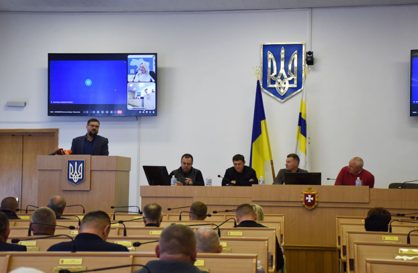 Засідання під час якого присутні сидять за столом, один зі спікером стоїть біля трибуни та говорить