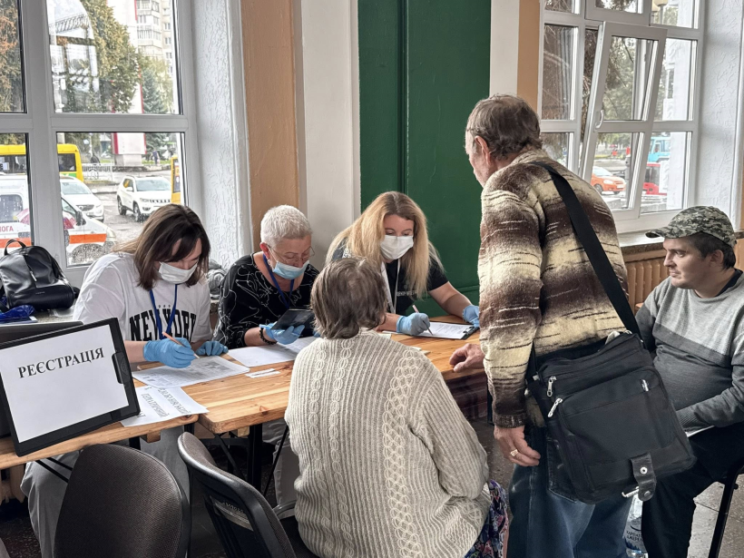 В залі очікувань за столом сидять представники місцевої влади та записують потреби ВПО. Біля столу сидять та стоять обпершись дві старші жінки