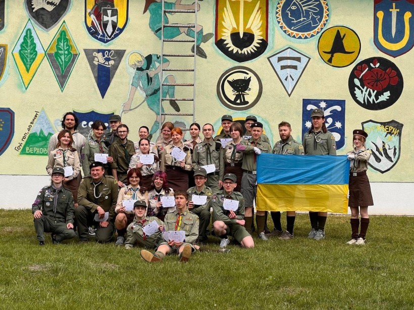 Загальне фото великої групи молоді-пластунів. Вониу сволїй формі та тримають у руках прапор України. Позаду них стилізована стіна, де розміщені крупним планом шеврони різних бойових бригад ЗСУ