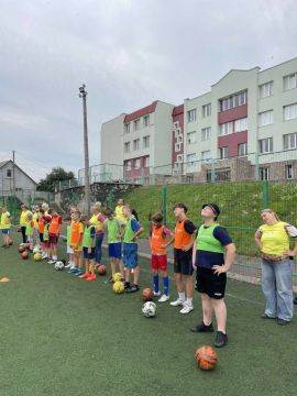 На спортивному майданчику біля школи  у шеренгу вистроїлися діти, підлітки та жінки. Вони проводять розминку тримаючи руки на талії та роблячи оберти головою.