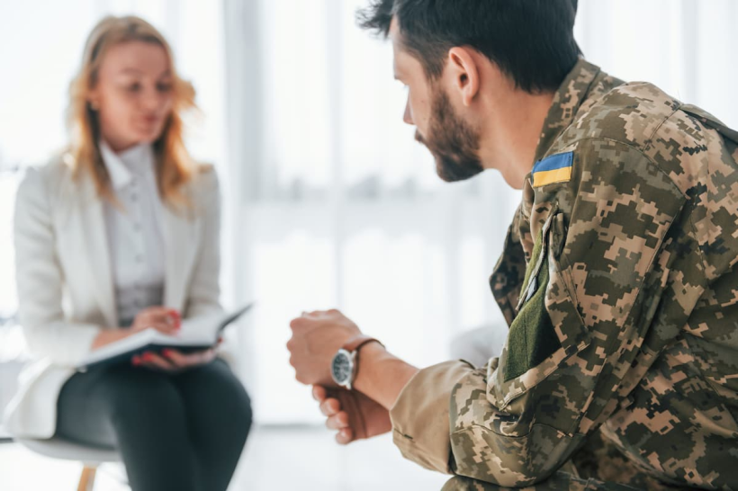 Фото ілюстративне - на передньому плані військовий. Він сидить напівбоком та спілкується з лікаркою, яка сидить на задньому плані та щось записує