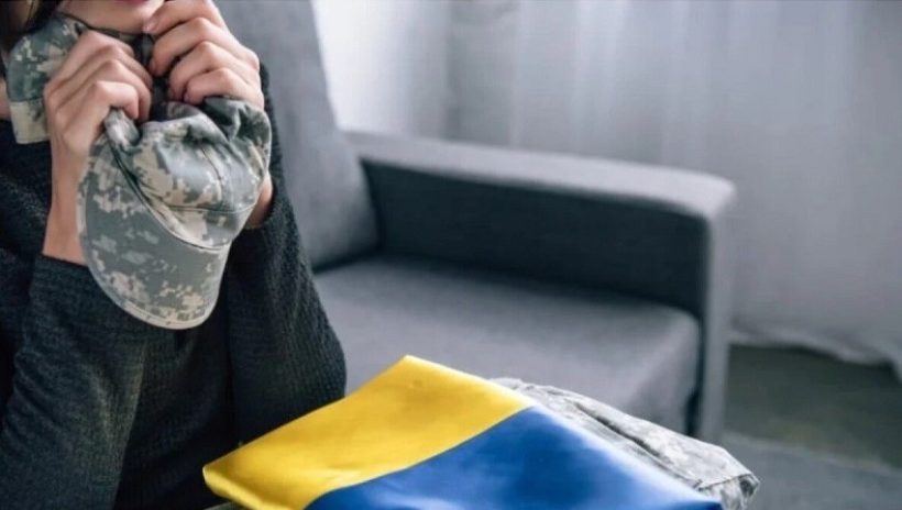 Фото ілюстративне: у кімнаті сидить жінка і тримає у руках піксельну кепку, а на колінах лежить прапор України