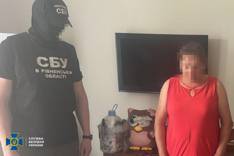 На фото зображено співробітника Служби безпеки України (СБУ) в Рівненській області та жінку, обличчя яких пікселізовані для збереження конфіденційності. Співробітник СБУ одягнений у чорну футболку з написом «СБУ в Рівненській області» та кепку з аналогічним маркуванням. На задньому плані видно телевізор, м’яку іграшку у вигляді сови та велику пластикову пляшку, обгорнуту чимось схожим на поліетилен чи тканину. Жінка стоїть поруч, одягнена у червоний топ без рукавів. У нижньому лівому куті зображений логотип Служби безпеки України.

Фото має офіційний характер і, ймовірно, зроблене під час проведення оперативних дій.