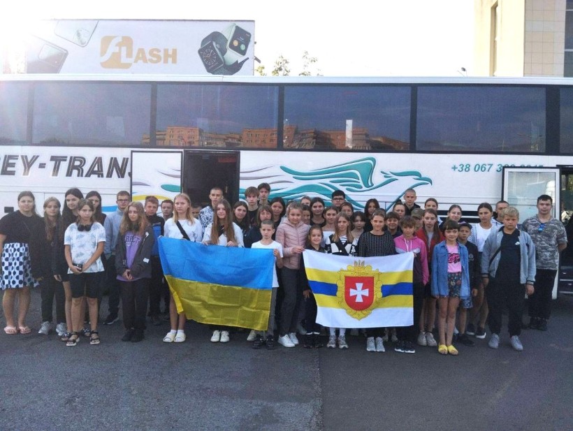 Діти, які їдуть на оздоровлення, стоять біля автобуса і тримають в руках Державний Проапор України та прапор Рівненської області