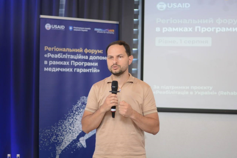 Начальник Рівненської ОВА Олександр Коваль під час форуму звертається до присутніх учасників зустрічі. В нього у руках мікрофон і він говорить. Позаду банер на якому написано - Регіональний форум "Ребілітаційна допомога в рамках програми медичних гарантій"
