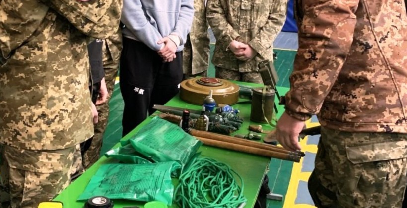 Люди у військовому та цивільному одязі стоять біля столу, на якому лежать елементи військового обладнання