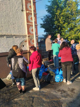 На світлині зображено групу людей біля будівлі під час гуманітарної допомоги. Це переселенці з Дніпропетровщини, які отримують необхідні речі та продукти. Люди стоять поруч із пакунками в синіх і чорних пакетах з речами першої необхідності. Дехто спілкується, дехто очікує своєї черги.
