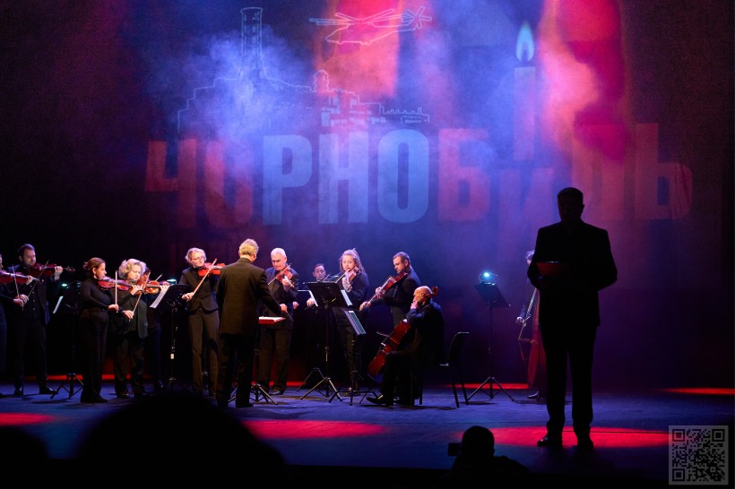 На фото зображена театральна сцена під час мистецького заходу. У центрі — камерний оркестр зі струнними інструментами, яким диригує чоловік у чорному костюмі. Музиканти зосереджено виконують твір, сидячи та стоячи біля пюпітрів із нотами.

Праворуч видно силует ведучого або читця з папкою в руках, який стоїть у напівтемряві. Сцену огортає густий дим і драматичне синьо-червоне освітлення, що створює напружену атмосферу.

На задньому плані великими літерами написано слово «Чорнобиль», а також проглядаються символічні зображення атомної станції, свічки та гелікоптера. Це свідчить, що подія присвячена темі Чорнобильської трагедії, пам’яті її жертв або героїзму ліквідаторів.

Загальна атмосфера фото — урочиста, емоційна та пам’ятна.