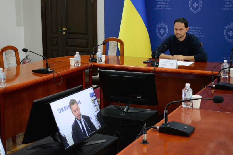 Начальник Рівненської ОВА Олександр Коваль сидить за столом та уважно дивиться у монітор, на якому заступник Міністра розвитку громад та  територій України Олексій Рябикін проводить онлайн-нараду