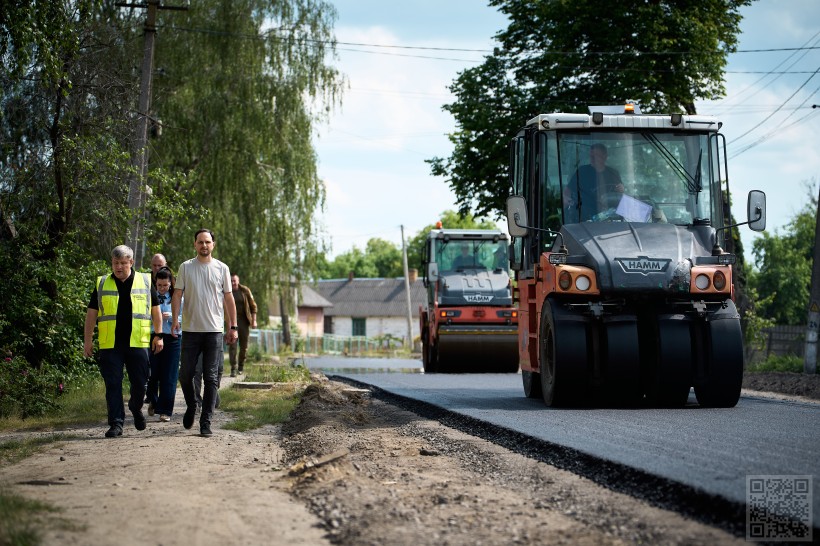 На автодорозі у селі спецтехніка укладає асфальт, поруч узбіччям іде начальник Рівненської ОВА Олександр Коваль та спілкується із будівельниками. Чоловіки одягнуті у жовті жилетки, розповідають очільнику області про хід ремонту автодороги