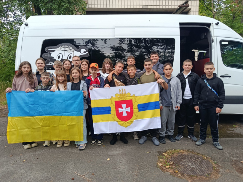 Група дітей стоїть біля автобуса. Вони посміхаються та тримають у руках жовто-блакитний прапор та прапор Рівненщини